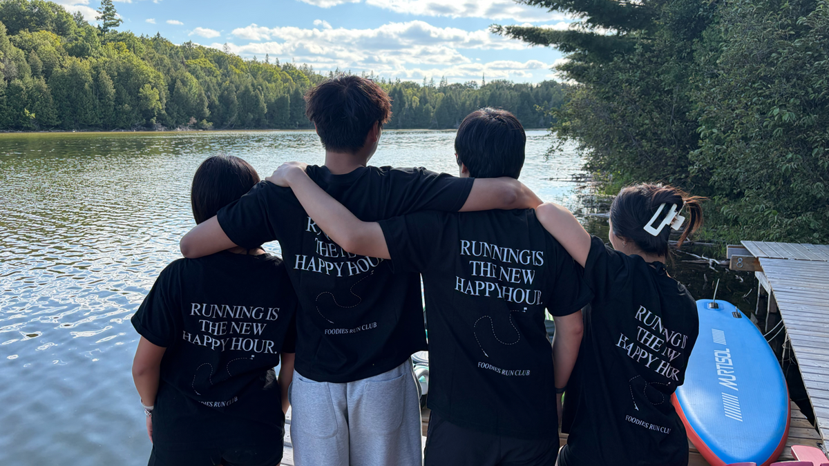Four people standing on a wooden dock by a lake, seen from behind, wearing matching black t-shirts with white text that reads 'RUNNING IS THE NEW HAPPY HOUR' and 'CHAMPSONIER'.